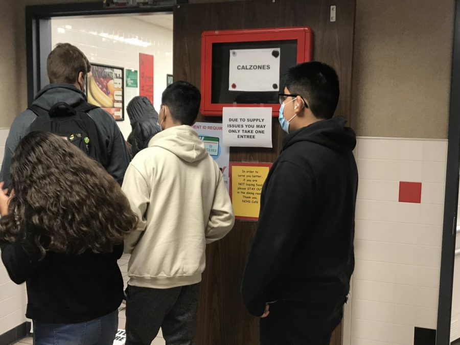 Students stand in line at lunch waiting to pick up their food. Signs hang that let students know they are only allowed to take one entree. 
