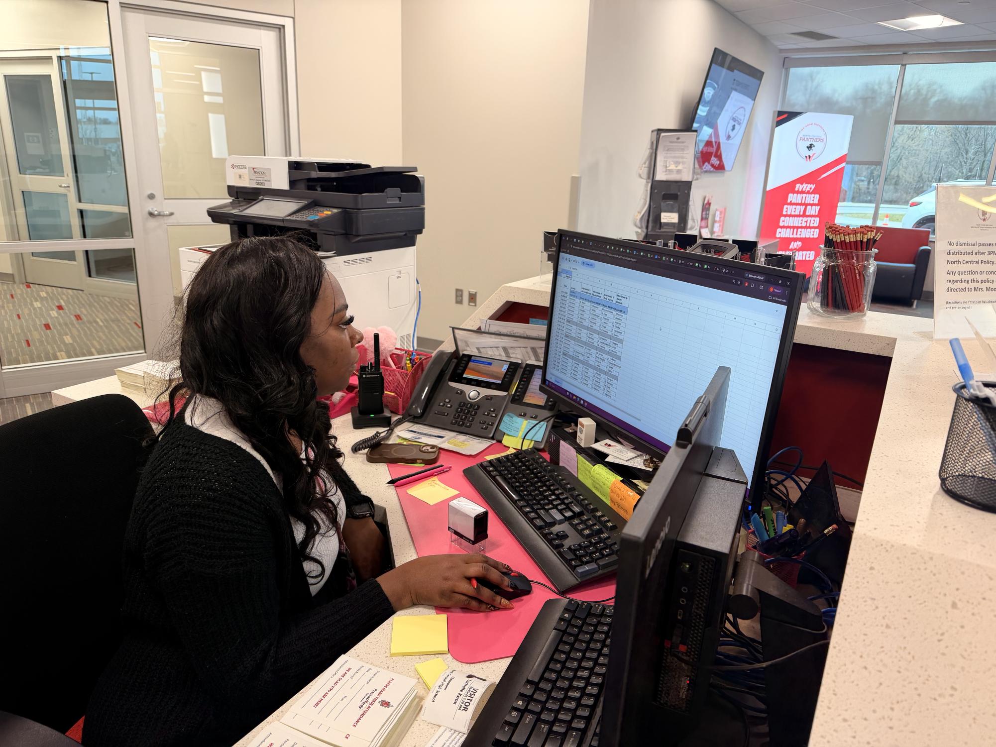 Jasmine Green greets students and helps guests from behind the front desk at North Central High School. Green said her job involved interacting with hundreds of students and parents throughout the day. She wanted to give students “something happy to see and not someone yelling at them,” Jasmine Green said.